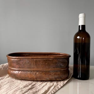 Può includere: Un vaso per piante ovale in rame martellato con un distinto bordo orizzontale che corre attorno al centro. Il rame ha una calda patina marrone metallizzato. Una bottiglia di vino in vetro marrone scuro si trova accanto al contenitore.
