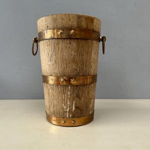 Könnte beinhalten: Ein rustikaler Holzeimer mit Metallbändern und Griffen. Der Eimer besteht aus hellfarbenem Holz mit sichtbarer Maserung und einigen Stellen mit abblätternder Farbe. Die Metallbänder sind kupferfarben, die Griffe sind rund.