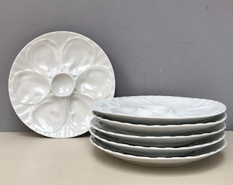 May include: A set of six white porcelain oyster plates. The plates are round and have six individual indentations for serving oysters. The plates are stacked on top of each other.