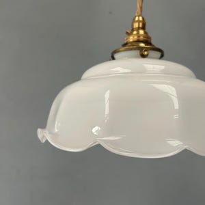 May include: A white glass pendant light shade with a scalloped edge and a smooth, glossy finish. The shade is attached to a metallic brass-colored fixture at the top, which connects to a light-colored fabric cord for hanging from a ceiling.