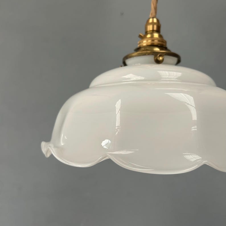 May include: A white glass pendant light shade with a scalloped edge and a smooth, glossy finish. The shade is attached to a metallic brass-colored fixture at the top, which connects to a light-colored fabric cord for hanging from a ceiling.