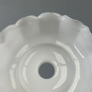 May include: A white glass lamp shade featuring a smooth, glossy finish and a scalloped, ruffled edge. The shade has a circular opening at the center, designed for a light fixture attachment, with a wide, bowl-like shape and curved sides.