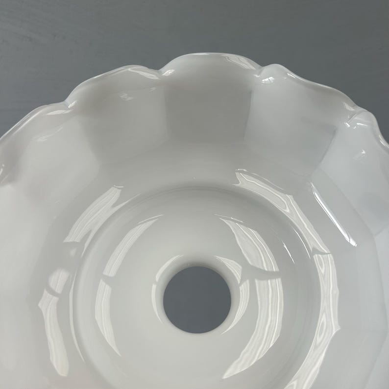 May include: A white glass lamp shade featuring a smooth, glossy finish and a scalloped, ruffled edge. The shade has a circular opening at the center, designed for a light fixture attachment, with a wide, bowl-like shape and curved sides.