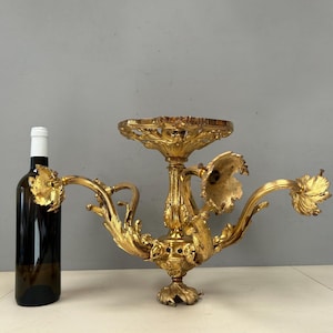 Könnte beinhalten: Vergoldeter Kerzenhalter mit drei geschwungenen Armen und einer zentralen Plattform. Der Kerzenhalter weist aufwendige Blumen- und Blattmotive auf. Daneben steht eine dunkle Glasflasche mit weißem Etikett.