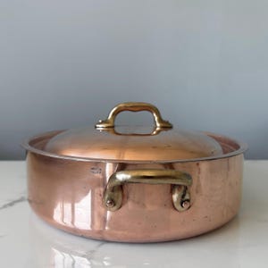 May include: A round copper cooking pot with a matching lid, featuring two brass side handles and a brass lid handle. The side of the pot is stamped with the text MADE IN FRANCE. The pot has a polished metallic finish and a low-profile design.