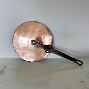 May include: A copper-colored, round pan lid with a long, dark handle. The lid has a slightly concave shape and a riveted handle. The handle is dark brown and has a loop at the end. The letters "I D" are stamped on the lid.