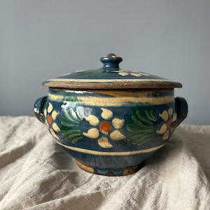 May include: A round ceramic lidded bowl featuring a deep blue glaze with hand-painted cream and green floral motifs. The pot has two side handles, a matching lid with a central knob, and a visible crackled finish across the entire surface.
