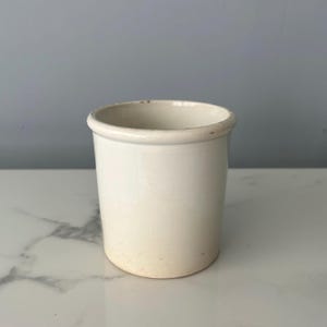 May include: A cream-colored ceramic jar with a rounded rim. The cylindrical container has a slightly aged appearance, with subtle discoloration near the base. The jar is set against a neutral background, highlighting its simple, classic design.