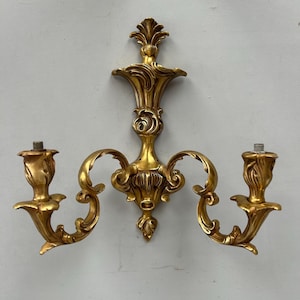 Könnte beinhalten: Vergoldete Wandleuchte mit zwei Kerzenhaltern. Die Leuchte hat ein dekoratives, symmetrisches Design mit geschwungenen Armen und floralen Akzenten. Die Leuchte ist aus Metall gefertigt und hat eine glänzende Oberfläche. Die Kerzenhalter sind für Standardkerzen konzipiert.