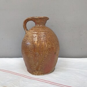 May include: A brown ceramic pitcher with a textured surface and a handle. The pitcher has a slightly rounded shape and a wide opening.