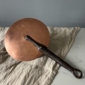 May include: A circular copper lid featuring a dark, textured iron handle attached with two rivets. The handle extends from the center to the edge and terminates in a circular loop at the end. The copper surface shows natural variations in color.