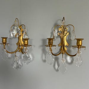 May include: A pair of gold-toned metal wall sconces, each featuring two candle holders and hanging clear glass crystals. The sconces have a central decorative backplate with curved arms extending to support the candle cups and dangling crystal accents.