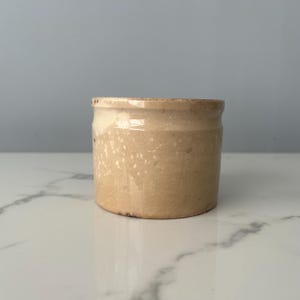 May include: A small, round, beige stoneware jar with a slightly textured surface. The jar has a cylindrical shape with a slightly wider opening and a subtle, light-colored rim. The surface has small, irregular white speckles.