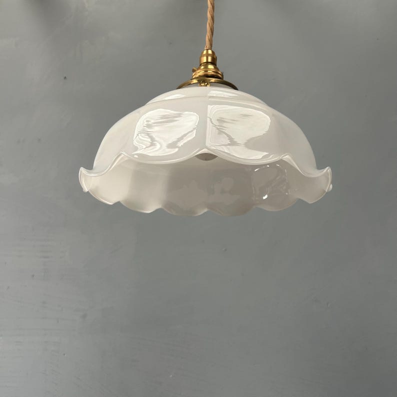 May include: A white glass pendant light fixture featuring a scalloped, ruffled edge design. The shade is attached to a brass-colored metal fitting and hangs from a twisted, light-colored fabric cord, creating a classic, clean lighting element.