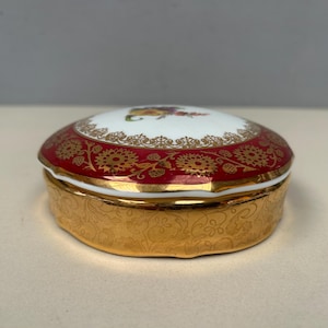 May include: A small, round trinket box with a white lid and a floral design. The lid has a red border with gold floral patterns. The base is gold with a floral pattern. The box is likely made of porcelain or ceramic.