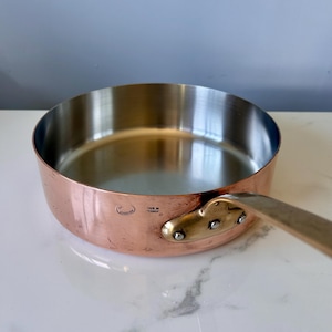 Könnte beinhalten: Eine runde Kupfersautépfanne mit einer polierten, silberfarbenen Innenauskleidung. Die Außenseite hat ein warmes Kupferfinish mit einem Messinggriffansatz, der mit drei Nieten befestigt ist. Der eingeprägte Text an der Seite lautet Made in France.
