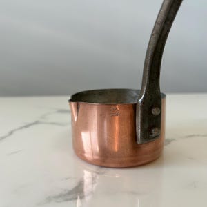 May include: A small copper saucepan with a riveted metal handle. The pot has a shiny, reflective surface and a spout for pouring. The handle is attached with rivets. The pot has a logo stamped on the side.