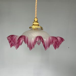 Puede incluir: Lámpara colgante de estilo vintage con una pantalla de cristal con volantes y borde rosa. La pantalla tiene un diseño esmerilado y arremolinado y está suspendida por un accesorio dorado y un cable trenzado beige. Decoración del hogar.