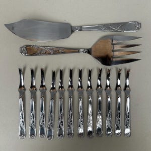 May include: A set of silver-colored serving utensils, including a fish knife, a three-tine fork, and twelve small forks. The handles feature an ornate floral design. The utensils are arranged on a light gray surface.