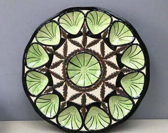 May include: A white ceramic plate with a black rim and green and brown leaf design. The plate has 12 individual compartments, each shaped like a scallop shell.