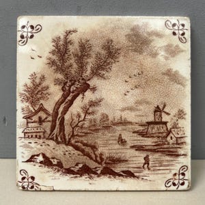 May include: Square ceramic tile featuring a sepia-toned landscape. The image shows a tree, buildings, a windmill, and figures near water. Decorative floral corner accents frame the scene.