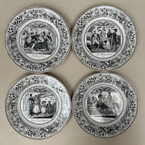 May include: A set of four antique plates, each with a black and white illustration. The plates have a floral border and a central hexagonal image depicting scenes. The plates are vintage and decorative, with detailed designs.