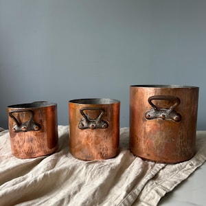 May include: A set of three cylindrical copper pots of varying sizes, each featuring a hammered texture and two dark, weathered metal handles attached with three visible rivets. The pots have a warm, metallic finish and a simple, functional design.