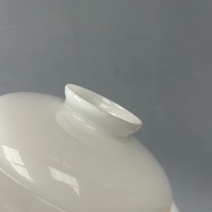 May include: A smooth, white, opaque glass lamp shade featuring a rounded, dome-like shape with a flared, circular opening at the top. The surface of the glass is glossy and reflects light, showing a clean, uniform finish throughout the entire piece.