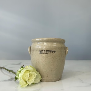 May include: A beige stoneware crock with the word "BILLETES" printed in black. The crock has two small handles and a slightly flared rim. A yellow rose lies in front of the crock.