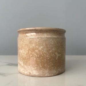 May include: A small, cylindrical stoneware jar with a speckled, light brown and white finish. The jar has a slightly flared rim and a smooth, unglazed surface. The overall aesthetic is rustic and vintage.