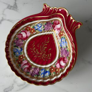May include: A shell-shaped porcelain dish featuring a deep red border with gold trim. The center is decorated with a gold floral motif, surrounded by a white band and a colorful painted wreath of roses, tulips, and small blue and orange flowers.