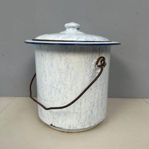 Puede incluir: Un cubo de esmalte blanco vintage con tapa, con un patrón azul veteado. Tiene un asa de metal marrón oscuro y un borde azul. El cubo probablemente se utiliza para almacenamiento o fines decorativos.