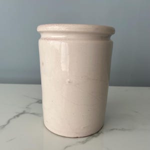 May include: A cylindrical, off-white ceramic jar with a slightly flared rim. The jar has a smooth, glossy finish with subtle variations in colour and texture. The rim has a reddish-brown hue. The jar is likely a vintage or antique piece.