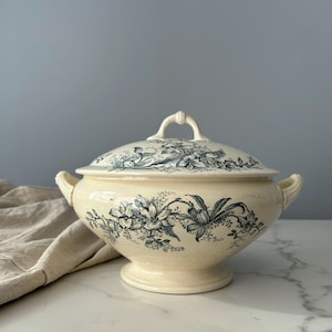 May include: A cream-coloured ceramic tureen with a matching lid, featuring a dark green floral transferware pattern. The tureen has two side handles, a pedestal base, and a decorative handle on the lid, showcasing detailed botanical illustrations.