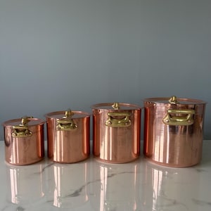 Op de afbeelding: Vier koperen potten van verschillende formaten staan op een rij. Elke pot heeft een deksel en messing handvatten. Het koper heeft een glanzend, reflecterend oppervlak. De potten staan op een wit marmeren oppervlak.