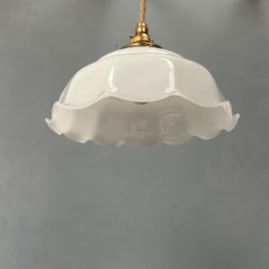 May include: A white glass pendant light fixture featuring a scalloped, ruffled edge shade. The lamp is suspended by a twisted beige fabric cord and includes a polished brass metal fitting at the top where the shade connects to the cord.