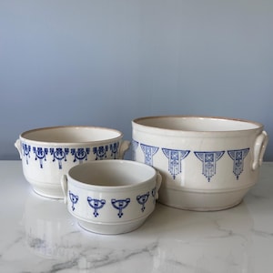 May include: Three white ceramic bowls with blue decorative patterns. The bowls vary in size, with the largest having a diameter of approximately 25 cm. Each bowl has small handles and a blue geometric design around the rim.