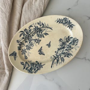 Könnte beinhalten: Eine ovale Keramikplatte mit cremefarbener Basis, die ein dunkelblaues Transferdruckmuster aus botanischen Blumensträußen, blättrigen Ranken und drei Schmetterlingen zeigt, die über die Oberfläche des Geschirrs verteilt sind.