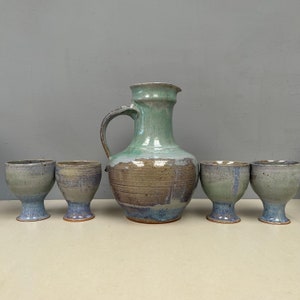 Könnte beinhalten: Ein Set aus fünf Keramikstücken, bestehend aus einer Kanne und vier Kelchen. Die Kanne ist hellgrün mit braunen und blauen Akzenten. Die Kelche sind in einem ähnlichen hellgrünen Farbton mit braunen und blauen Akzenten gehalten. Die Stücke sind alle handgefertigt und haben ein rustikales, erdiges Aussehen.