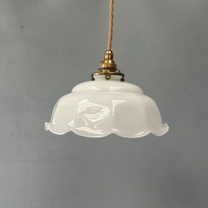 May include: A white glass pendant light fixture with a scalloped edge, featuring a brass-toned metal fitter and a twisted beige fabric cord. The shade has a smooth, opaque finish and a rounded, bell-like shape suspended from the ceiling fixture.