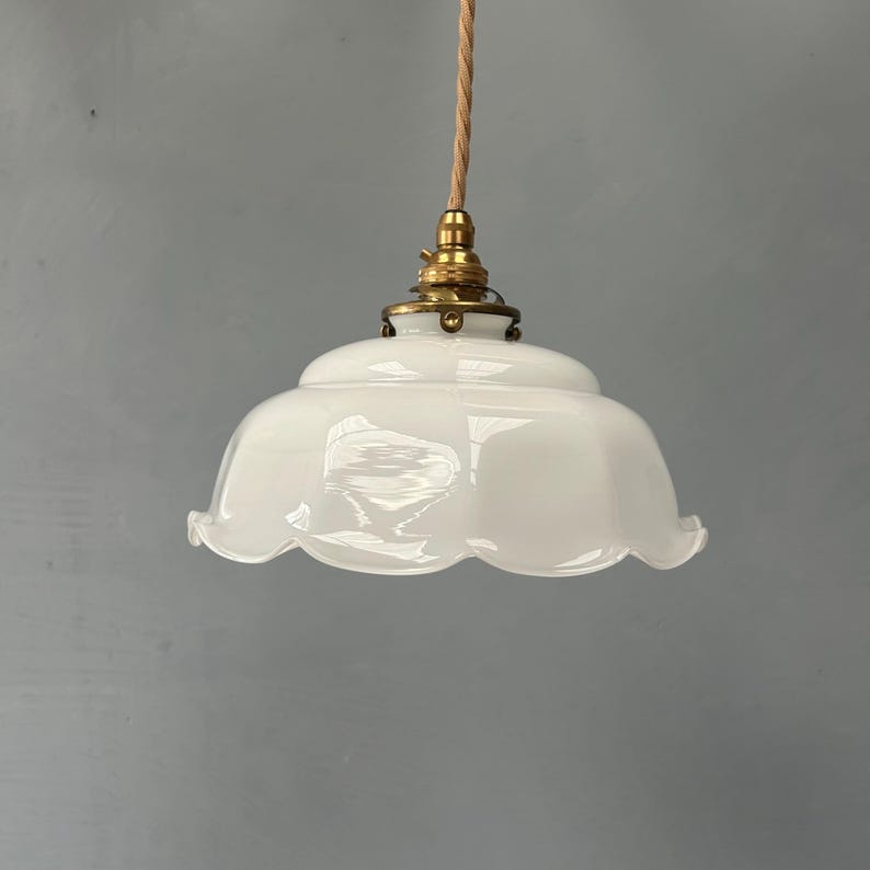 May include: A white glass pendant light fixture with a scalloped edge, featuring a brass-toned metal fitter and a twisted beige fabric cord. The shade has a smooth, opaque finish and a rounded, bell-like shape suspended from the ceiling fixture.