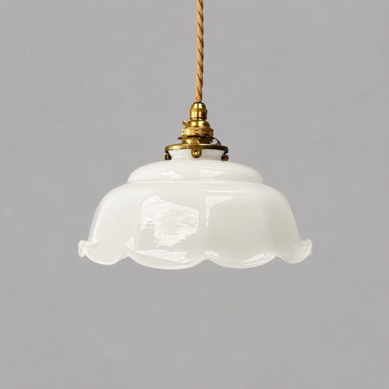 May include: A white glass pendant light fixture with a scalloped edge, featuring a brass-colored metal socket and a twisted tan fabric cord. The shade has a smooth, glossy finish and a wide, rounded shape that flares outward at the bottom rim.