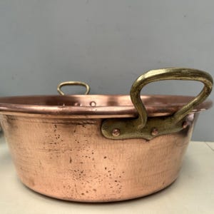 Peut inclure: Une marmite en cuivre avec deux poignées en laiton. Le pot a une texture martelée et une forme arrondie. Les poignées sont incurvées et fixées au pot avec des rivets. Le pot est probablement utilisé pour la cuisine ou le service.