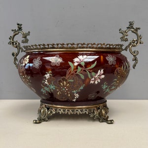 May include: A large, oval-shaped ceramic bowl with a red base and floral pattern in shades of pink, white, green, and gold. The bowl has ornate brass handles and a base with three feet.