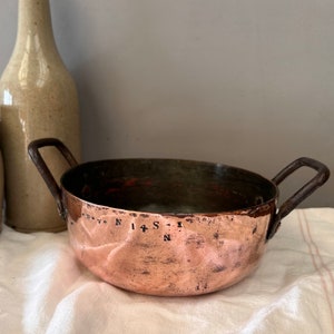 May include: A copper pot with two handles and a hammered finish. The pot has a dark copper color and is inscribed with the text "D.V.N.I 4 S.I N".