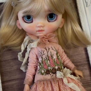 Può includere: Una bambola d'arte personalizzata con capelli biondi e occhi blu brillanti. La bambola indossa un vestito rosa con dettagli floreali ricamati e una gonna di pizzo. Un delicato fiocco adorna l'abito. Il viso della bambola ha un incarnato morbido e rosato.