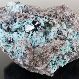May include: A close-up of a rock with a blue-green mineral coating. There are several small, black, shiny crystals embedded in the rock.