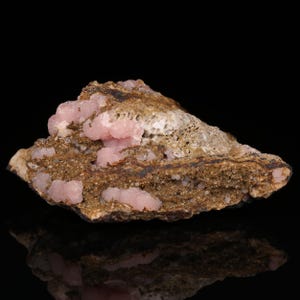 Puede incluir: Un grupo de cristales de rodocrosita rosa incrustados en una matriz marrón. Los cristales son pequeños y tienen una forma redondeada. La matriz es rugosa y texturizada.