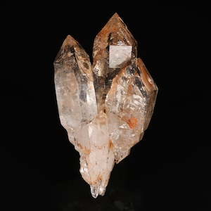 May include: A cluster of clear quartz crystals with amber and orange hues. The crystals have sharp, pointed tips and a rough, textured base. The quartz is translucent, allowing light to pass through, against a black background.