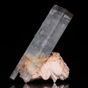 Puede incluir: Un gran cristal transparente de color azul claro con estrías en su superficie. El cristal descansa sobre un grupo de cristales blancos más pequeños.
